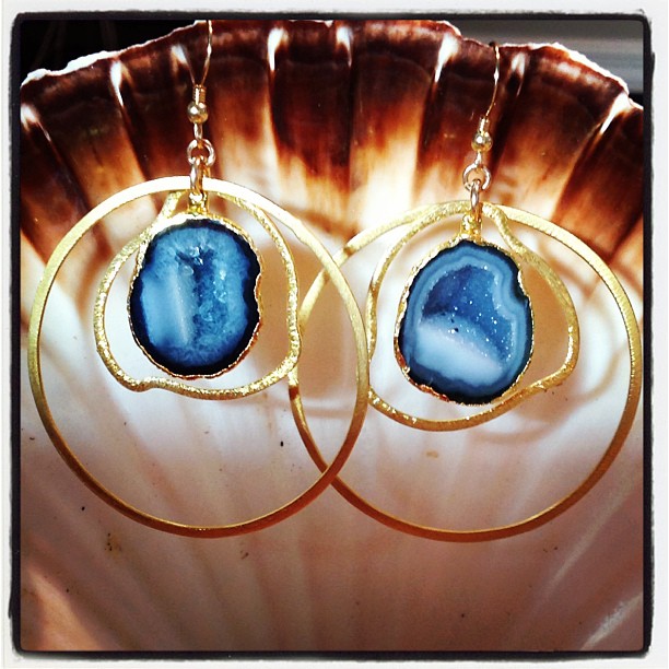 Blue Geode Hoops, featuring gold hoops and irregular blue geode stones, are shown hanging in front of a brown and white seashell to emphasize their textured, organic style.
