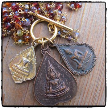 A close-up of the Buddha Amulet Necklace on Garnet Chain, featuring engraved Buddhist figures and Thai script on amulets attached to a gold keyring, displayed beside red, orange, and purple beads on a wooden surface.