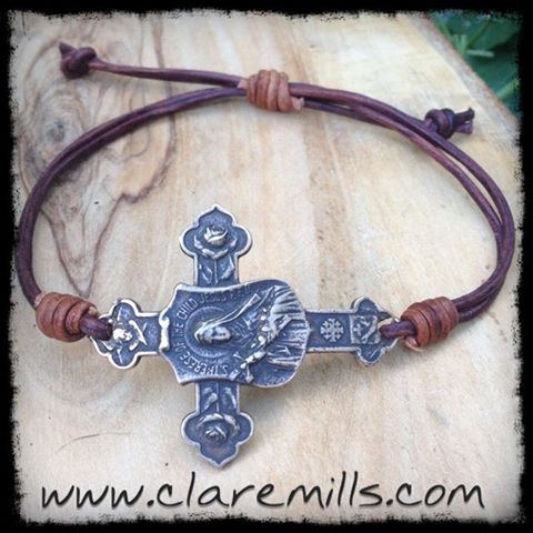 The St. Theresa Little Flower Cross on Leather bracelet features a brown leather cord with adjustable knots and an engraved silver cross. The text www.claremills.com appears at the bottom of the image.