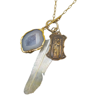 The “Guardian Feather” Geode & Mother-of-Pearl Toggle Necklace features a gold chain with three pendants: a white feather, a gold-edged geode slice, and an ornate gold saint charm.