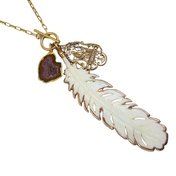 The “Crimson Guardian” Geode & Bone-Feather Toggle Necklace is a gold chain featuring a white feather, a gold-framed brown geode, and an ornate gold charm with intricate designs.