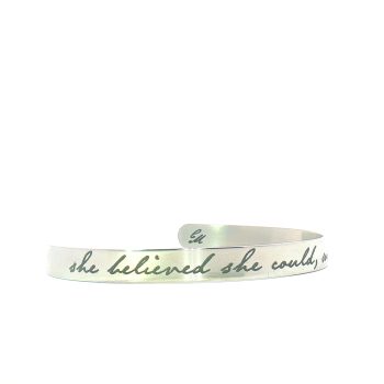 A silver cuff bracelet engraved with the phrase she believed she could in cursive script. The bracelet has an open, adjustable design and a brushed metal finish.