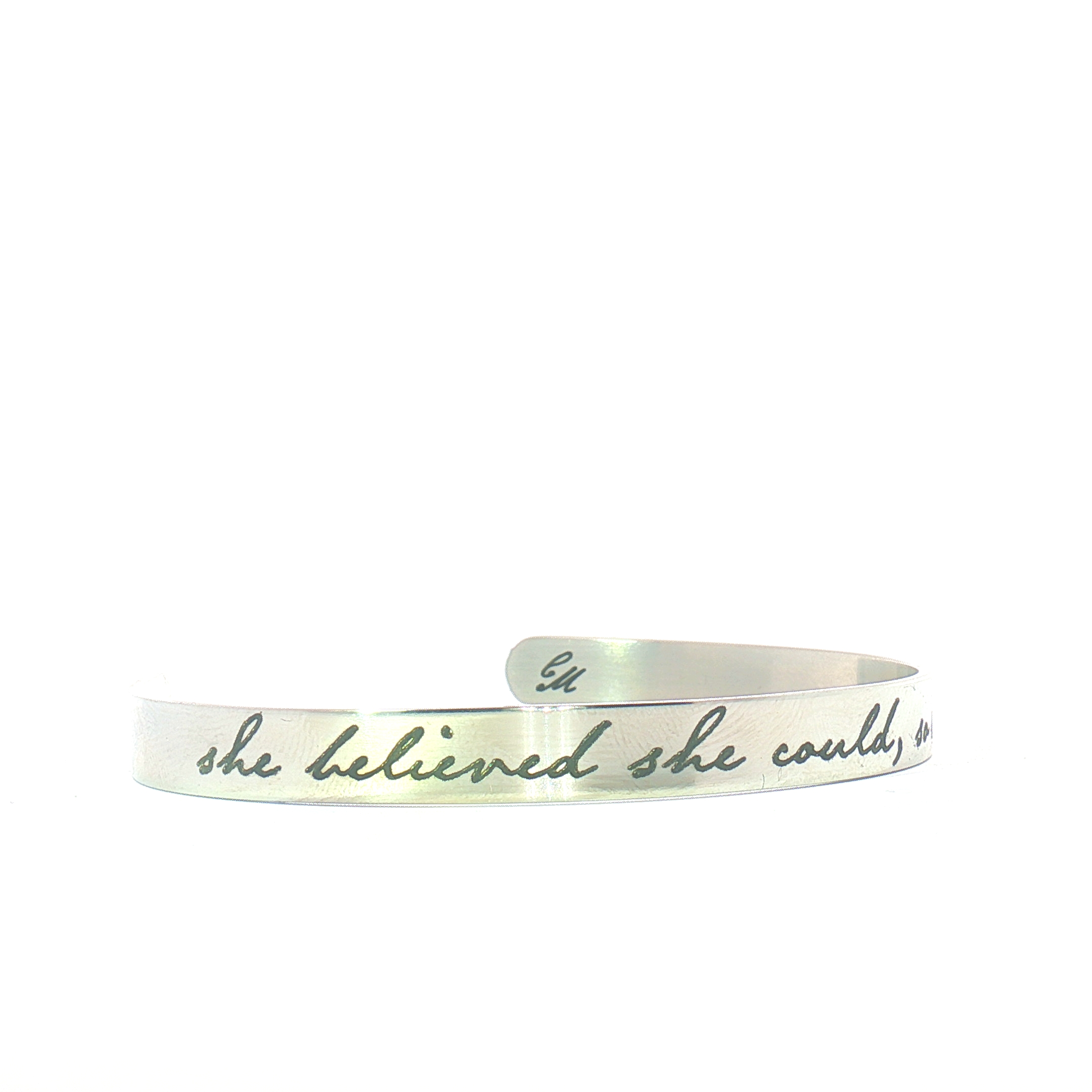 A silver cuff bracelet engraved with the phrase she believed she could in cursive script. The bracelet has an open, adjustable design and a brushed metal finish.
